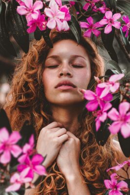 A serene woman with curly red hair surrounded by vivid pink blossoms, eyes closed.